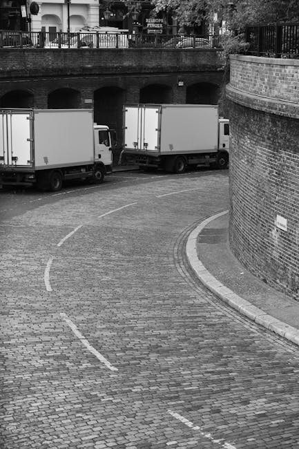 Black and white image showing a curved cobblestone street with two white moving vans parked alongside the curb. The vans are positioned in a line, facing away from the camera, and are in the process o
