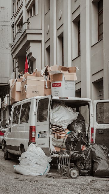 A white removal van parked on a city street beside a stone building with multiple windows and architectural details. The van's rear doors are open, revealing a cluttered interior filled with large pla