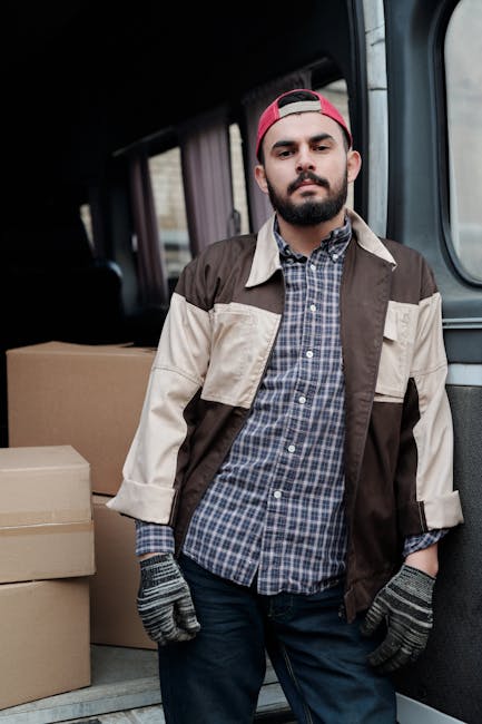 A man with a beard and wearing a red cap, a checkered shirt, a beige and brown jacket, and grey gloves stands inside a van during a home relocation. The van's interior shows cardboard boxes stacked on the floor behind him, some taped and sealed for transport. The man appears to be inspecting or preparing for the loading process, with his hands resting at his sides. The background includes part of the van's cargo area and a glimpse of the outdoors through the windows, indicating the ongoing process of furniture transport and packing involved in a house move. This setting illustrates typical steps in furniture transport and loading as part of a professional removals service, such as those provided by Man and Van Haringey, supporting efficient home relocation activities.
