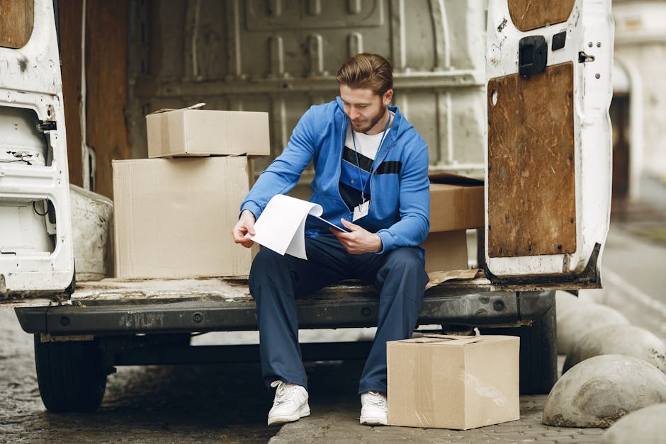 A man dressed in a blue jacket, white t-shirt, and dark trousers is sitting on the edge of an open moving van, surrounded by cardboard boxes of various sizes, some sealed with packing tape and others open, revealing packaging materials such as plastic wrap and packing paper. The man is holding a clipboard and pen, reviewing a checklist or inventory, likely related to home relocation or furniture transport. The interior of the van shows a partly visible wooden panel on the side, with additional boxes stacked inside. Outside, the van is parked on a paved surface with large concrete spheres nearby, indicating a loading zone. The scene captures the process of packing and organizing household items for a professional removals service, with equipment such as the trolley or straps not visible but implied, and the lighting indicates daylight, highlighting the logistical aspects of packing and moving furniture efficiently. Man and Van Haringey provides these moving and packing logistics support, focusing on proper handling and transportation of household belongings during house removals.