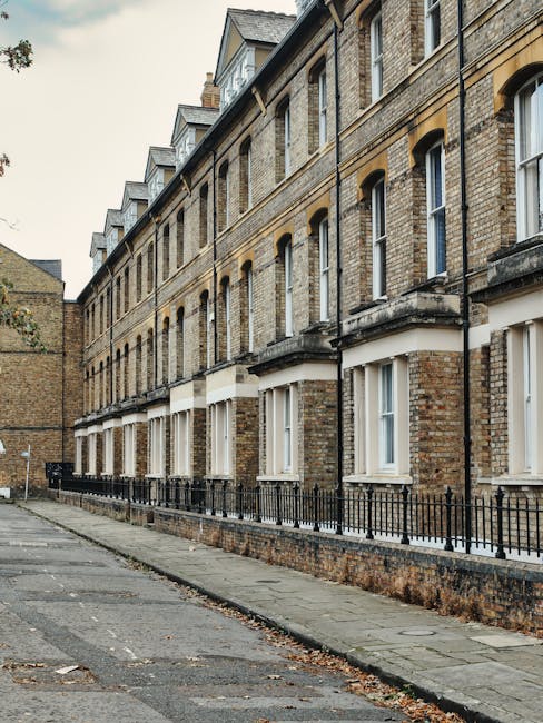 A narrow street adjacent to a multi-storey brick terraced house with several windows, some open with white frames, and a row of small dormer windows on the roof. The building is constructed of yellow-brown brick and features decorative stone lintels above the windows. A black metal railing runs along the low brick wall at pavement level, separating the property from the sidewalk. The pavement appears slightly uneven with scattered leaves and debris, indicating an outdoor residential area. No moving activity or equipment is visible in this image. This setting exemplifies typical urban housing often encountered in house removals and relocation services in Haringey, where narrow access routes may present challenges during furniture transport and home relocation tasks managed by Man and Van Haringey.