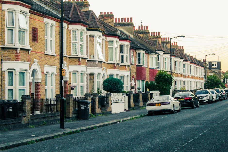A row of terraced residential houses in Haringey with brick facades, some featuring decorative white window frames and bay windows, situated along a quiet street during daylight. The pavement is lined with small fences and gardens, and several parked cars, including a white sedan, are visible along the curb. In the foreground, a man and van vehicle operated by Man and Van Haringey is actively engaged in a home relocation process, with people lifting or carrying cardboard boxes and furniture wrapped in plastic or blankets out of a property and onto the pavement for loading into the van. The scene captures the logistics of packing and furniture transport during a house move, with an emphasis on the practical aspects of narrow access removals, illustrating coordination in loading household items through potentially restricted access points. Street lamps and overhead power lines create a typical urban neighbourhood environment, emphasizing the challenges and solutions associated with moving in residential areas with limited access.