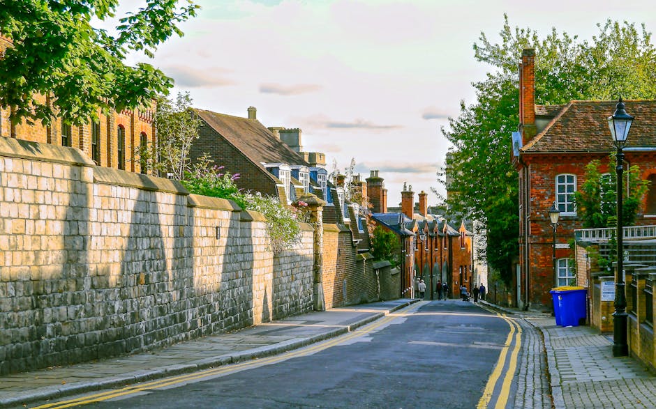 A narrow residential street in Crouch End, Haringey, featuring a gently sloping tarmac road with double yellow lines along the curb. On the left side, a high stone wall separates the sidewalk from brick and stone houses with pitched roofs and chimneys, some with small front gardens and flowering plants. On the right, there are older brick buildings with white-framed windows, street lamps, and a blue recycling bin next to the pavement. In the distance, a few pedestrians are walking along the sidewalk, and a team from Man and Van Haringey is likely involved in home relocation or furniture transport services, as the scene suggests a typical urban setting suitable for loading and moving household items during a residential move.