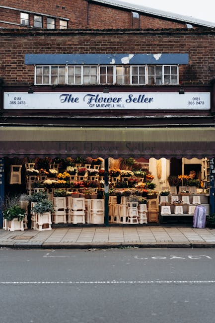 The storefront of 'The Flower Seller' shop located on a pavement, featuring a variety of fresh flowers and plants displayed on white wooden stands and tables inside the shop. The display includes bouquets, potted plants, and floral arrangements, with some flowers wrapped in paper or plastic. The shop has a brown awning above the entrance, and the signage shows the business name, phone number, and location as Muswell Hill. Above the shop, there is a brick building with a row of windows, and a blue metal sign with peeling paint is attached above the shopfront. The scene captures a typical high street shopfront during daytime, with natural light illuminating the floral display, which may relate to home relocation and packing services when considering moving or transporting delicate items like plants during a house move. Man and Van Haringey services offer assist with these types of relocation logistics, including furniture transport and careful handling of fragile items.