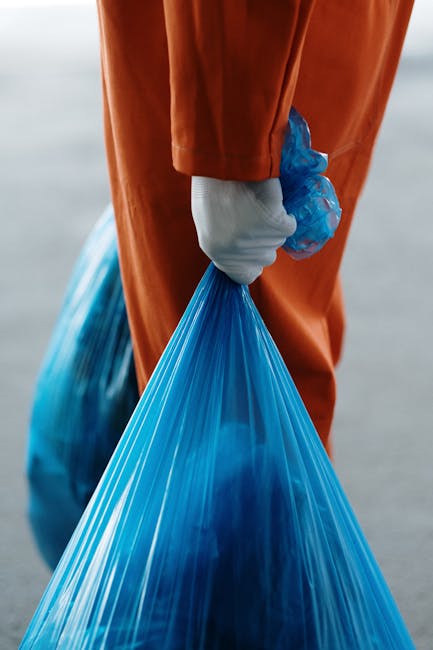 A person wearing orange overalls and a white glove is holding a large blue plastic garbage bag filled with waste, with their hand gripping the top of the bag. The background appears to be an outdoor u