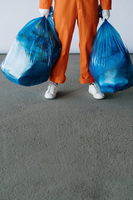 A person wearing orange overalls and a white glove is holding a large blue plastic garbage bag filled with waste, with their hand gripping the top of the bag. The background appears to be an outdoor u