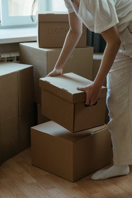 A person wearing light clothing and standing indoors, lifting a small cardboard box with their right hand. Several larger cardboard boxes are stacked on the wooden floor around them, some with labels such as 'CLOTH' written in red marker. Natural light illuminates the scene through a nearby window, casting soft shadows. The person appears to be in the process of packing or unpacking as part of a home relocation, with the boxes likely containing household items to be moved. This image illustrates the packing stage of a house removal, which is an integral part of furniture transport and moving logistics. The setting suggests a residential room with a clear focus on the careful handling of boxes, supported by the presence of furniture and packing materials, aligning with professional removals services such as those offered by Man and Van Haringey.
