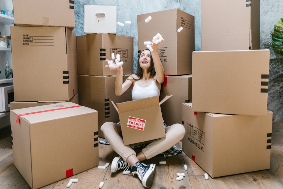 Two women are smiling and standing inside a property surrounded by packed cardboard boxes, some sealed with red tape, and a large potted plant with broad green leaves. One woman holds a small box, while the other carries a larger box and has her arm around her companion. They are positioned near a wall with a textured, light grey surface. The scene is well-lit, indicating indoor lighting suitable for a home relocation or moving process. Equipment such as packing materials and the presence of multiple boxes suggest they are in the midst of packing or unpacking during a house move. This setup is typical in a professional furniture transport or house removal scenario, with boxes arranged for easy loading or unloading, consistent with services provided by Man and Van Haringey. The environment emphasizes preparation for moving, with objects organized for efficient transport, supporting quick and smooth home relocations.